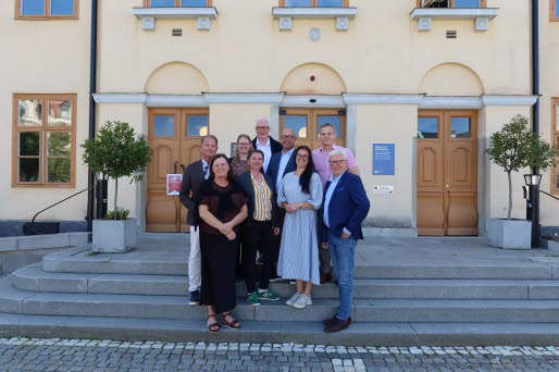 Män och kvinnor som är politiker eller tjänstepersoner i en kommun, står på en trappa framför ett gult hus