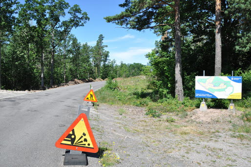 Byggarbete i Långelid. En stor skylt vid sidan av vägen berättar om att det kommer byggas nya villatomter i området. Även varningsskyltar för byggarbete syns vid vägen.