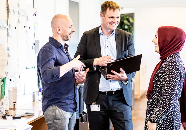 tre personerstår på ett kontor och samtalar. En av dem har en laptop öppen i händerna.