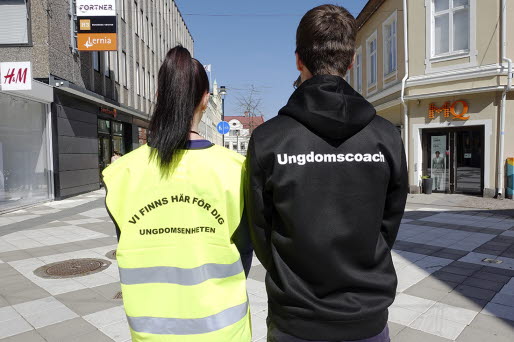Två personer vända med ryggen mot kameran. en person bär skyddsväst med texten Vi finns här för dig, ungdomsenheten. På den andra personens hoodie står det Ungdomscoach.