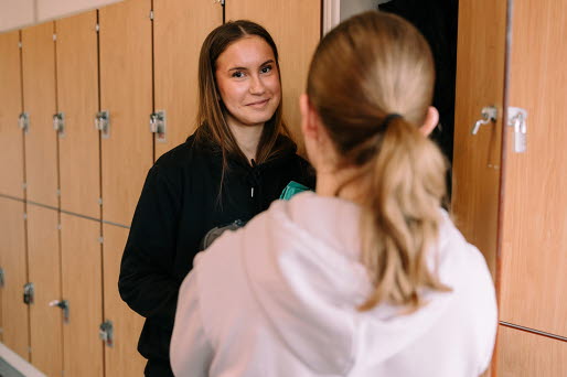 Två tjejer står vid sina skolskåp och pratar.