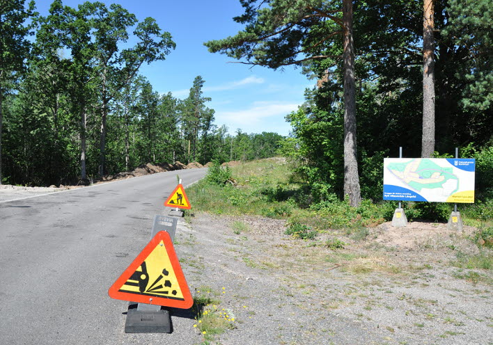 Byggarbete i Långelid. En stor skylt vid sidan av vägen berättar om att det kommer byggas nya villatomter i området. Även varningsskyltar för byggarbete syns vid vägen.