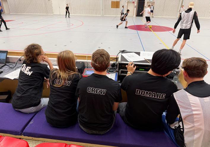 Fotbollsturnering inomhus. I förgrunden sitter några domare i bakgrunder ser man två lag spela fotboll.