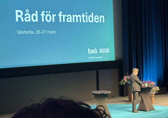 En person står på en stor scen och håller föredrag. I bakgrunden syns en Powerpoint-presentation.