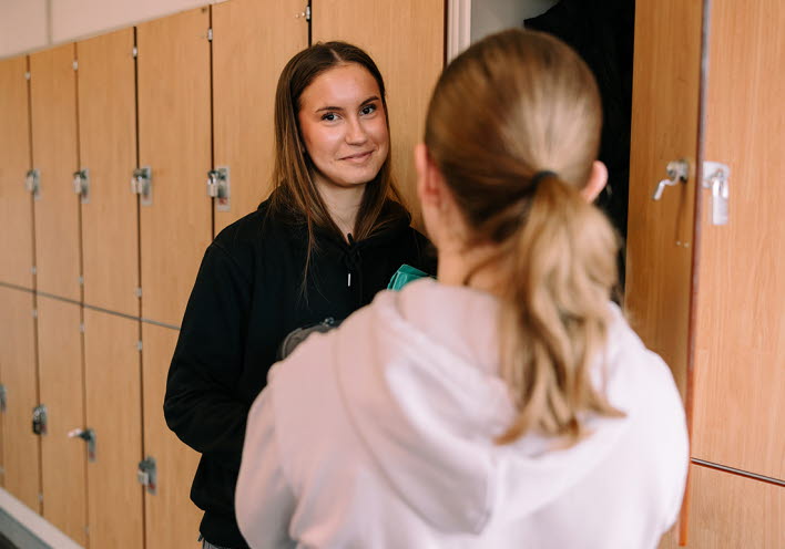 Två tjejer står vid sina skolskåp och pratar.