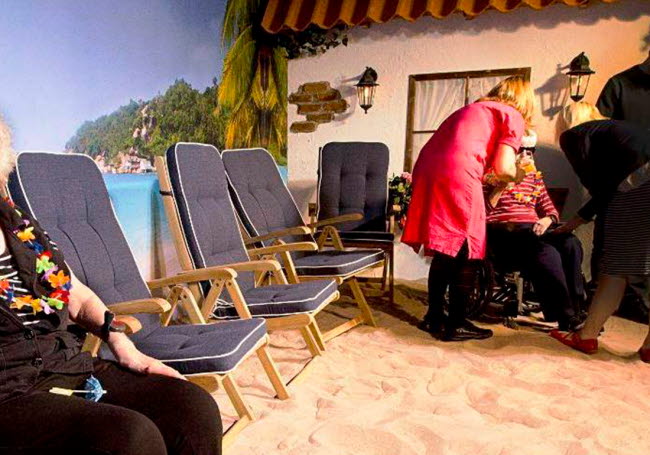 Några äldre personer sitter i solstolar på en konstgjord sandstrand med lampor som solljus.